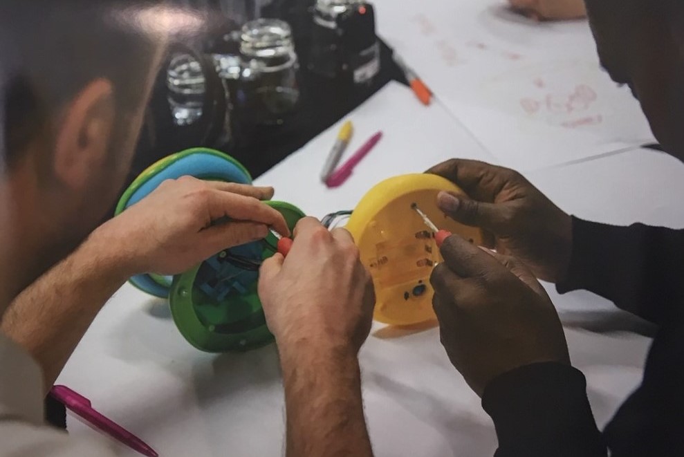 Toy autopsy in action: two sets of hands shown unscrewing toy parts on a tabletop